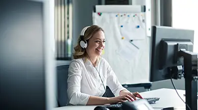 Applied Contact Centre Woman dressed in business casual attire smiling while sitting at desk using computer with headphones on.