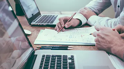 Ivans Exchange  Close up of two laptops and two colleagues interacting with stack of papers.