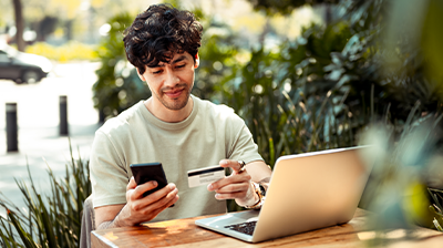Applied Pay A man sitting at a table outside, in front of an open laptop while holding his phone in one hand and a credit card in another hand