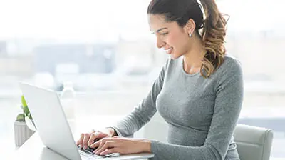 10 Email Best Practices for Sending Better Emails A woman in a grey sweater sitting at her desk smiling and typing on her laptop.
