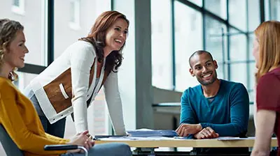 Latest Blog Post A woman leaning on a small table with three other people sitting down while all smiling at each other.