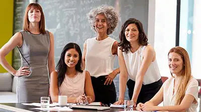 What Does Women’s History Month Mean to You? A group of five women dressed in business attire, standing and sitting around a table while smiling at the viewer