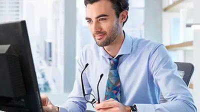 A man sitting in his office and looking at a computer screen while holding a pair of glasses in his hand