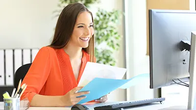 A woman holding files in her hands while smiling at her computer