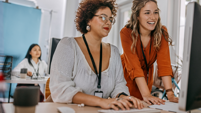 EZLynx Street Smart Webinar  Three women in a office at their desks. One woman is sitting at her desk using her computer while another woman in leaning on the desk and looking at the same computer