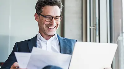 Industry Insight Comparative Quotes Man in business attire smiling while looking at laptop and holding documents.