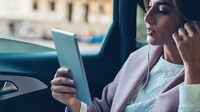 Insight The Cloud Woman in business attire sitting in car with window down using a tablet with earphones in her ears.