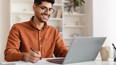An Up-Close Look at What Applied Marketing Automation Can Do A man in an orange shirt, sitting at a desk while using a laptop and holding a pen with one hand
