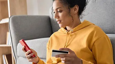 Your First Look – Applied Pay A woman in an orange hoodie using a red smart phone while holding a credit card with her other hand