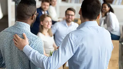 Producer Recruitment and Development A group of 5 coworkers sitting while looking at two men standing in front of them with their backs to the viewer. The man on the right is resting a hand on the other man's back.
