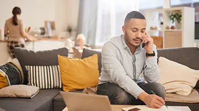 A man sitting on a couch and talking on his phone while taking notes. A woman is standing in the kitchen behind him