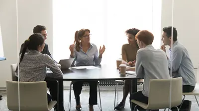 What It Takes to Be a Best Practices Agency Six people sitting around a table having a meeting. A woman in the middle is the main speaker while the others are listening.