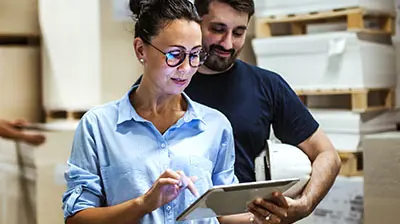 A woman using a tablet while a man holding a white hard hat is standing slightly behind her